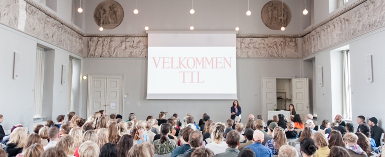 Mennesker, der sidder på rækker af stole rettet mod skærm på væg i Festsalen på Kunstakademiet