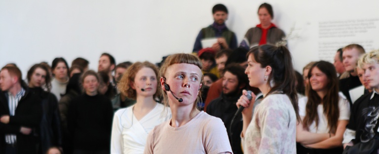 Tre personer med mikrofoner bliver betragtet af et publikum i forbindelse med performance