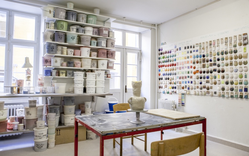 Bøtter med glasurblandinger på væghylder, brændte glasurprøver på væg og keramisk figur på drejeskive på bord i Laboratoriet for Keramik